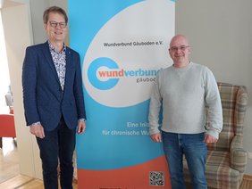 Zwei Herren vor einem blau-weißen Roll Up, Dr. Giesler im blauen Anzug, Herr Siebert mit Jeans und grauen Pulli