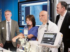 Vor dem großen Bildschirm und hinter dem Behandlungstisch mit Display, Kabeln, Infusionsflasche. Dr.. Günter in Kittel, Frau Kötterlin blauem Kasack, die anderen beiden Herren mit Sacko