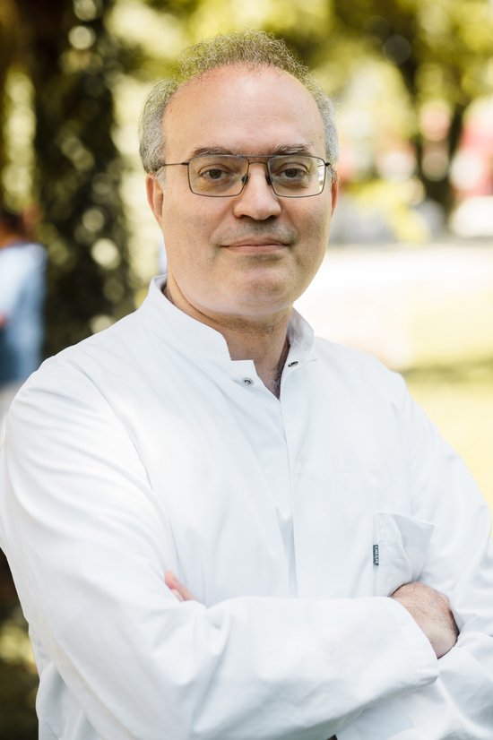 Gürol Salk (© Johannes Lehner) Portrait eines Arztes in Kittel mit verschränkten Armen, sympathisch lächelnd, Brille und grauem Haar mit Natur im Hintergrund