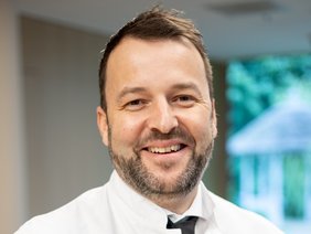 Prof. Dr. Thomas Betz, Chefarzt der Klinik für Gefäßchirurgie am Klinikum Straubing: „Im Vorfeld der Behandlung chronischer Wunden spielt die Gefäßdiagnostik zur Abklärung der Ursache eine wichtige Rolle.“ (© Johannes Lehner) Portrait von einem lachenden Arzt mit leicht melliertem Bart, Arztkittel und Krawatte