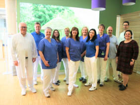 Team MVZ Strahlentherapie (© Sandra Wimmer) Die Gruppe steht vor einem hellgrünem LED-Bild mit Naturmotiv und auf hellem Holzboden, zwei lila Lampenschirme sieht man oben rechts. Die Ärzte sind in weißen Kitteln gekleidet, die anderen mit blauer Hose und dunkelblauem Polo, eine Dame in Zivilkleidung in Schwarz, rechts.