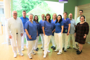 Team MVZ Strahlentherapie (© Sandra Wimmer) Die Gruppe steht vor einem hellgrünem LED-Bild mit Naturmotiv und auf hellem Holzboden, zwei lila Lampenschirme sieht man oben rechts. Die Ärzte sind in weißen Kitteln gekleidet, die anderen mit blauer Hose und dunkelblauem Polo, eine Dame in Zivilkleidung in Schwarz, rechts.