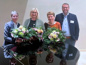 Alle vor einem Klavier, die Damen links mit großen Blumensträußen, zwei mit kurzen Haaren, alle lächelnd.