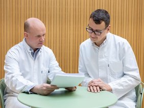 Zwei Ärzte an einem grünen Tisch in Besprechung. Der Herr mit wenig Haaren hält Dokumente. Der Herr rechts hat eine Brille und schaut interessiert darauf.