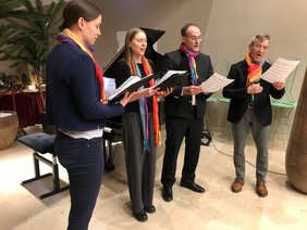 Links die Damen, alle dunkel gekleidet mit buntem Schal und Notenblätter in der Hand, singend. Die Herren mit Brille. Im Hintergrund Klavier und Krippe