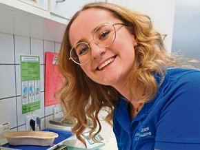Junge Frau in blauem Shirt mit halblangen blonden gewellten Haaren und Brille mit goldenem Gestellt sowie blauen Handschuhen und Glasplättchen vor sich lacht seitlich in die Kamera